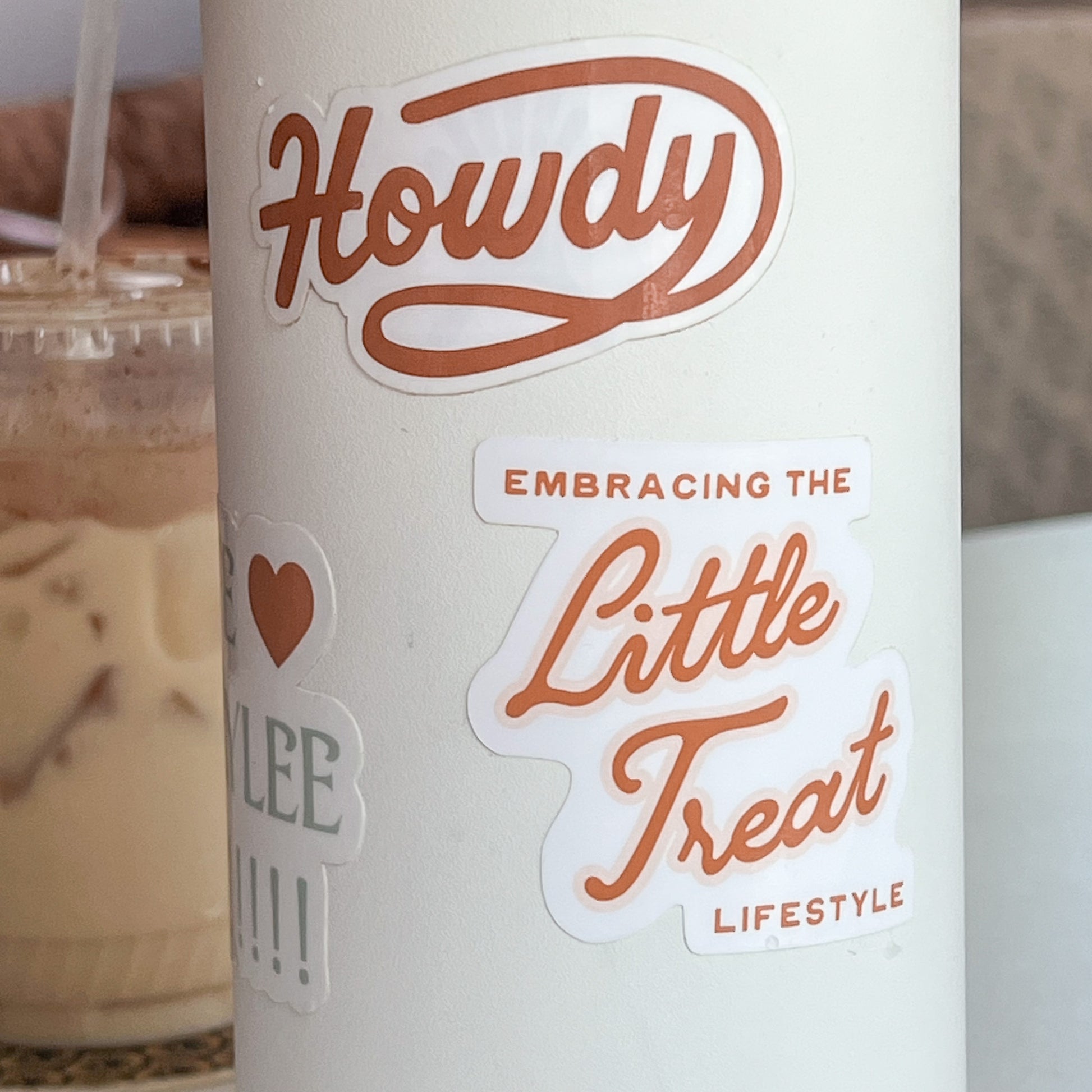 White Hydro Flask waterbottle featuring a sticker with the quote "Embracing the Little Treat Lifestyle" in pink and red. Iced Coffee is seen in the background