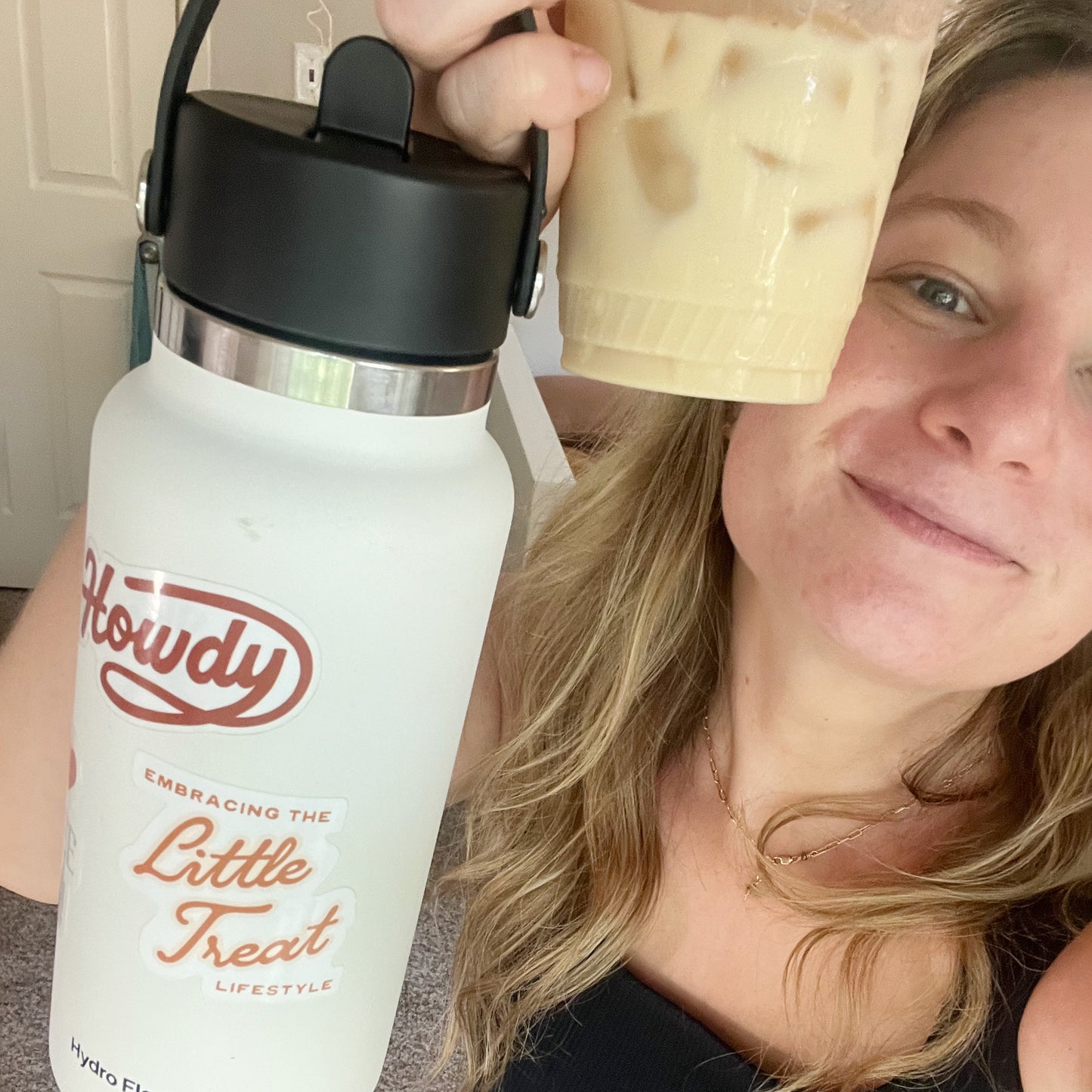 Selfie of a woman holding an iced coffee and a white hydro flask water bottle featuring a sticker with the quote "Embracing the Little Treat Lifestyle" in pink and red