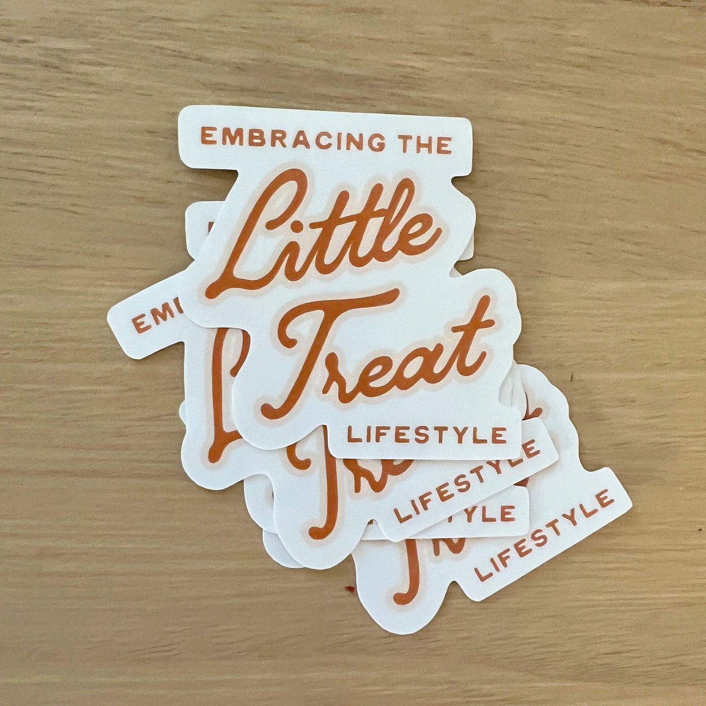 Light wood background featuring a pile of stickers with the quote "Embracing the Little Treat Lifestyle" in pink and red