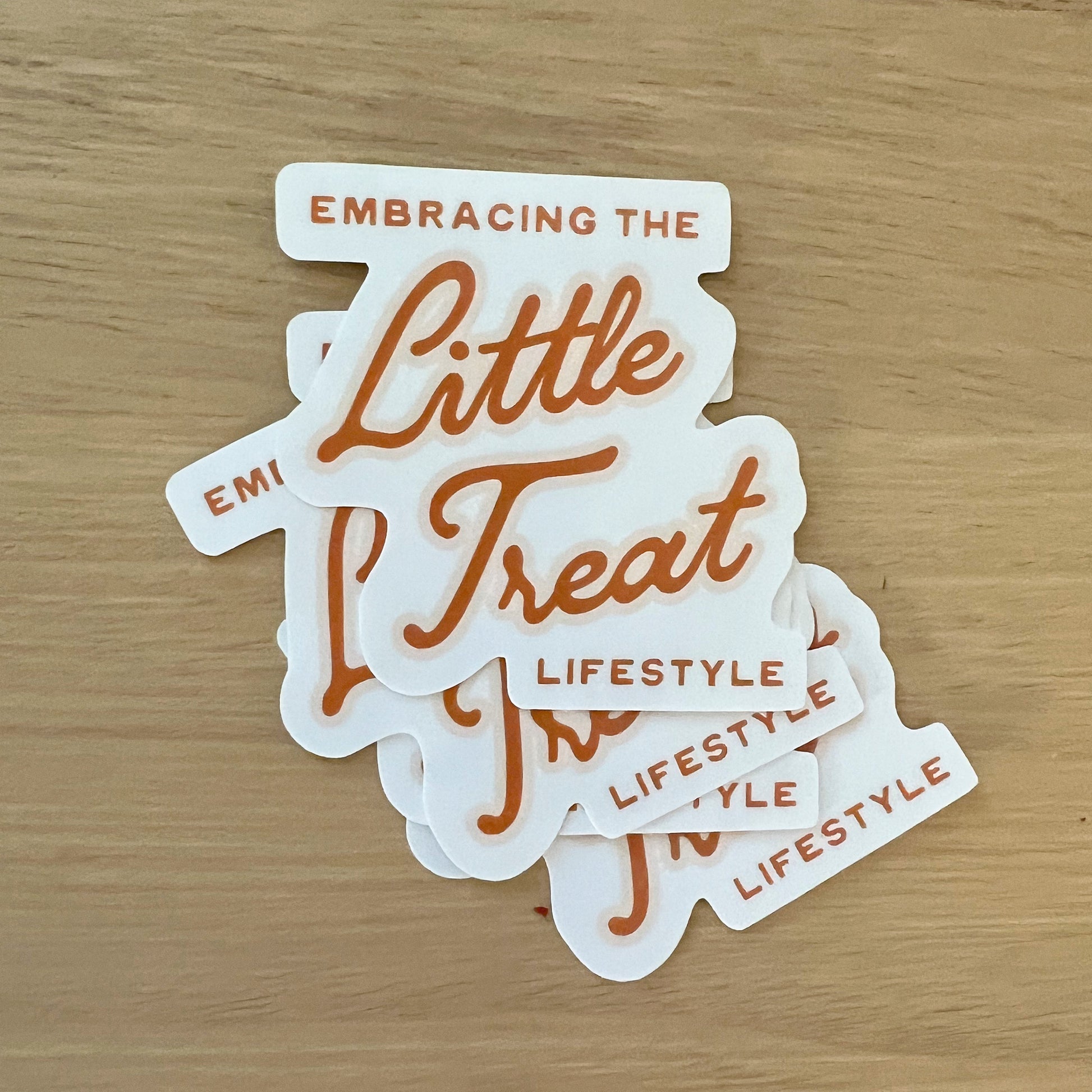 Light wood background featuring a pile of stickers with the quote "Embracing the Little Treat Lifestyle" in pink and red