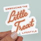 Blue background featuring a sticker with the quote "Embracing the Little Treat Lifestyle" in pink and red