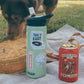 Camelbak water bottle with 3 stickers on it on a picnic blanket next to a dog, a canned cocktail, and a wicker purse