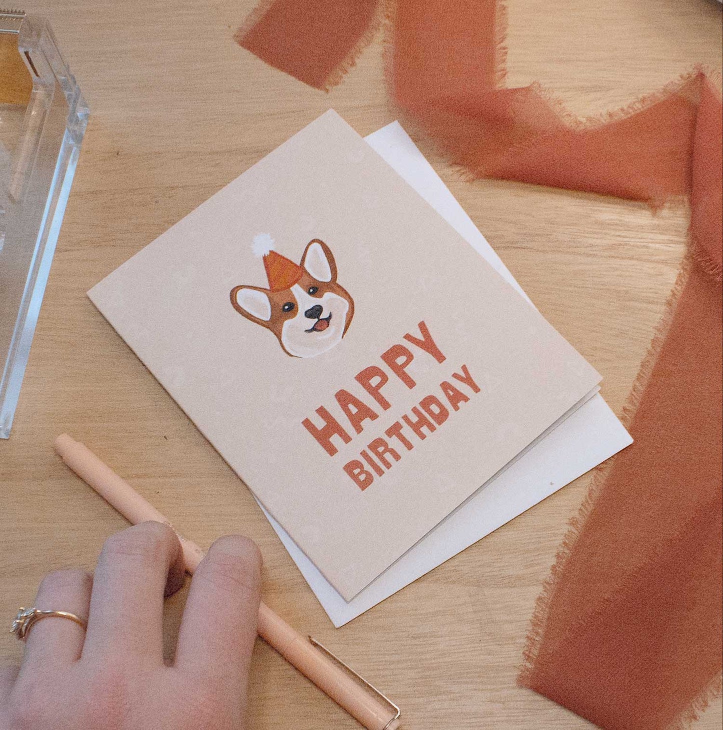 Corgi in birthday hat card on a light wood background with a hand, a rust colored chiffon ribbon, and a peach colored pen