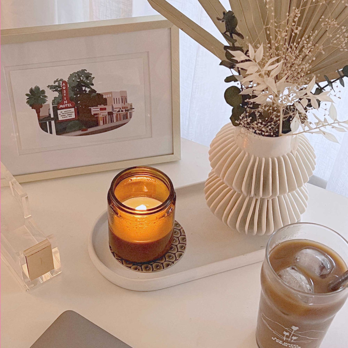 South Congress postcard in a gold frame next to a white, ribbed CB2 vase with dried florals, iced coffee, and a PF Candle Co. Candle